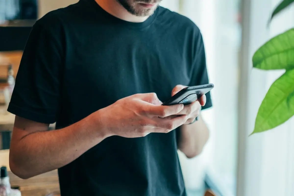 Homem jovem vestindo camiseta preta, segurando um celular smartphone e olhando para a tela, em um ambiente iluminado com plantas ao fundo. (Imagem: Jonas Leupe/Unsplash)
