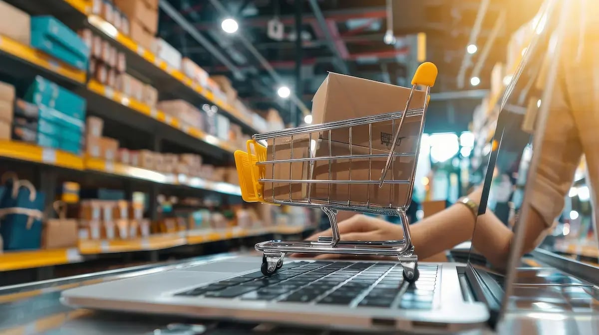 Carrinho de compras em miniatura com caixas de papelão apoiado no teclado de um notebook aberto, com prateleiras de um centro de distribuição ao fundo.