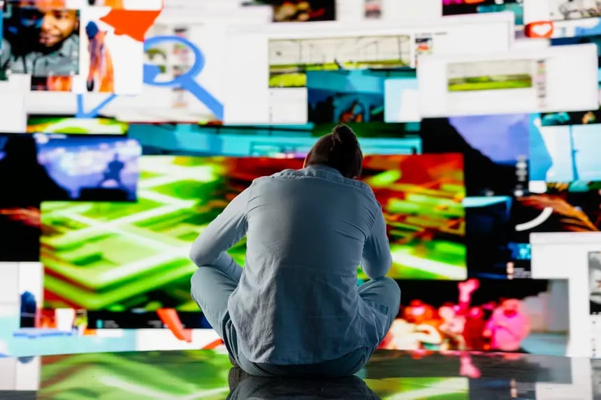 Pessoa sentada de costas diante de um mural de telas digitais conectadas, representando a expectativa para a chegada da TV 3.0 no Brasil a partir de 2026 e a convergência entre sinal aberto e internet.