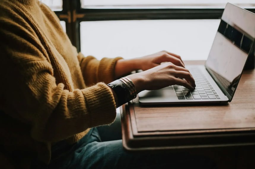 Pessoa usando um suéter amarelo digitando em um notebook prateado sobre uma mesa de madeira, com luz natural entrando pela janela ao fundo. (Imagem: Christin Hume/Unsplash)